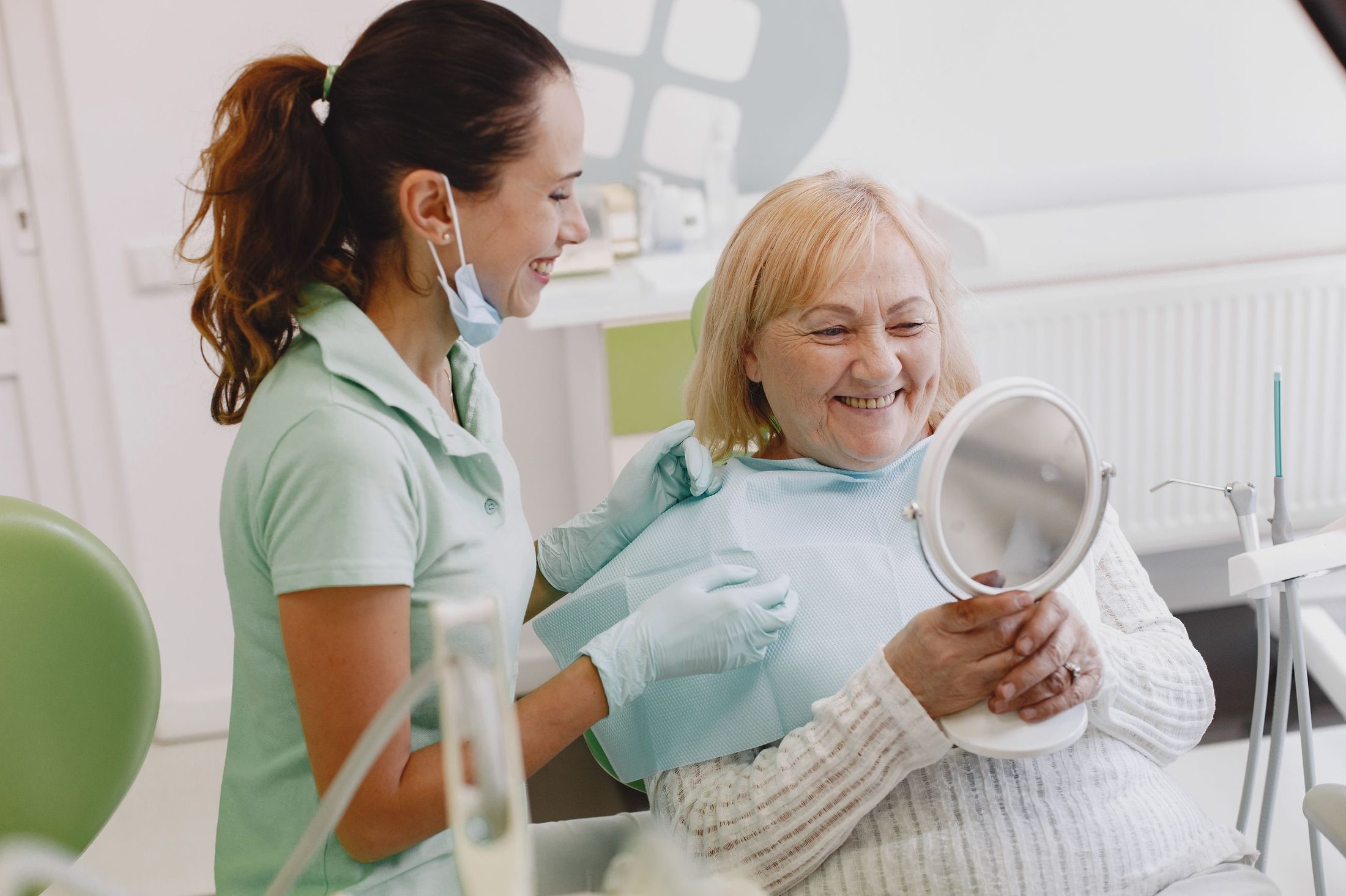 An older woman smiling at her new teeth in a hand mirror, with a dental assistant
